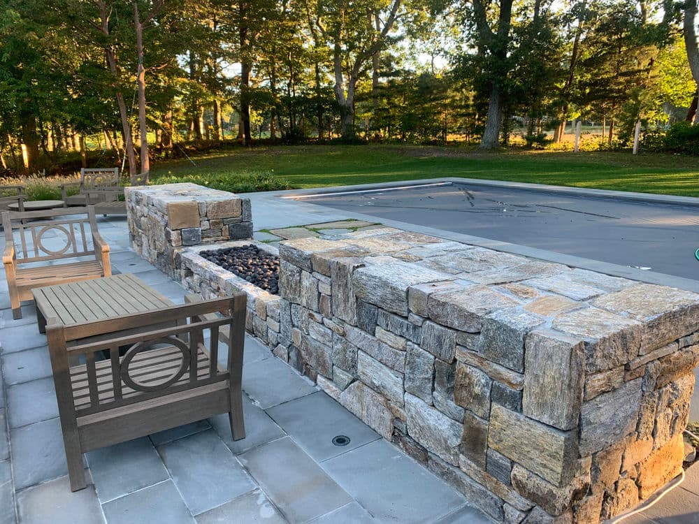 Stone fire pit with seating area beside a covered swimming pool in a landscaped yard.