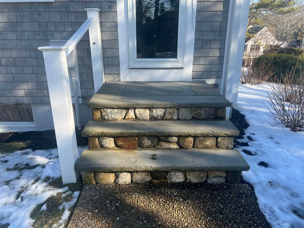 Stone steps leading to a home's front door, surrounded by snow and landscaping.