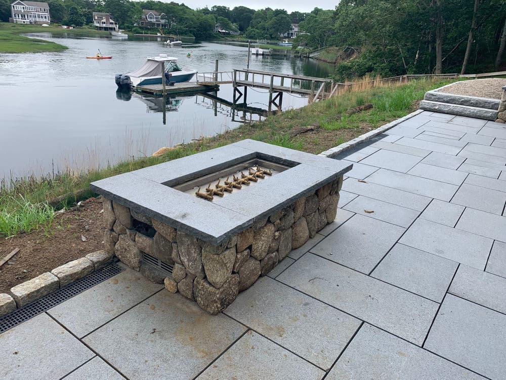 Stone fire pit by a serene waterfront, surrounded by lush greenery and wooden dock.