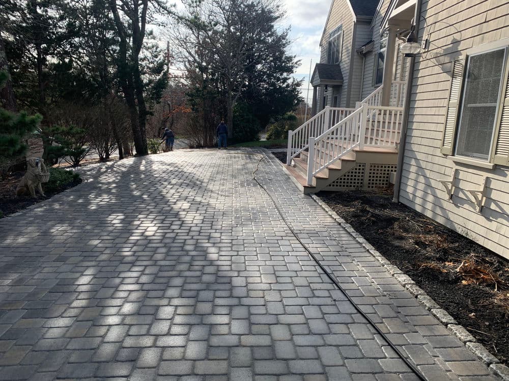 Paved driveway beside a house, featuring trees and a clear sky in the background.