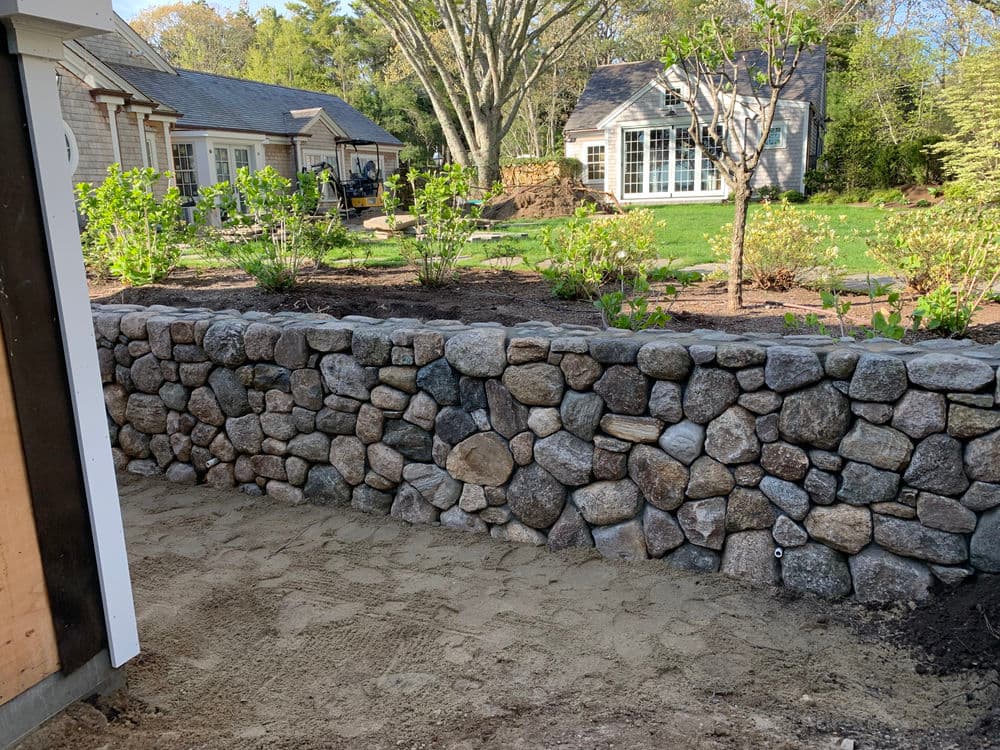 Stone wall in landscaped yard with new plants and trees, alongside a house.