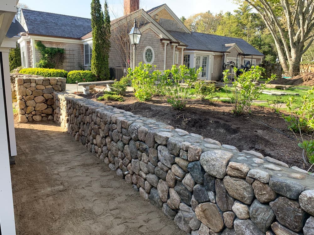 Stone wall along a landscaped pathway leading to a house with green plants and trees.