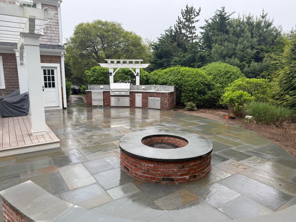 Outdoor patio featuring a stone fire pit, built-in grill, and lush greenery in the background.