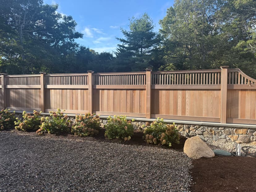 Wooden fence with decorative top, surrounded by gravel and shrubs under a blue sky.