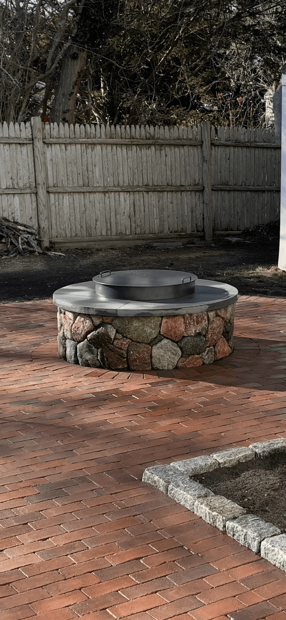 Stone fire pit on a brick patio surrounded by a wooden fence and trees.