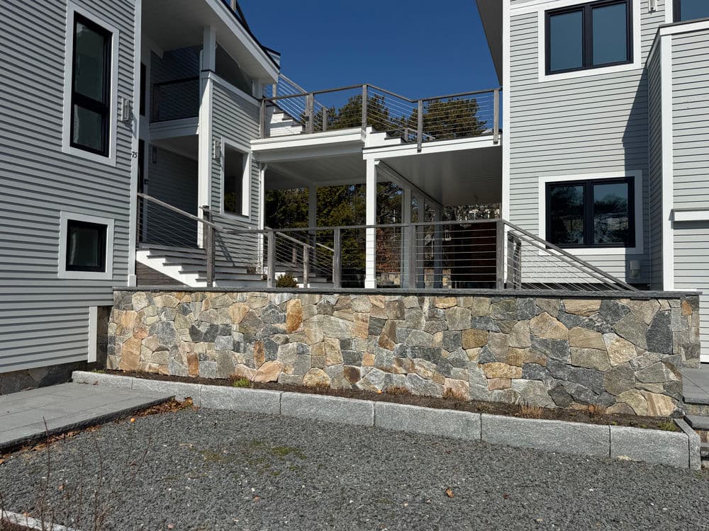 Modern house exterior featuring stone wall, deck, and paved pathway under clear blue sky.